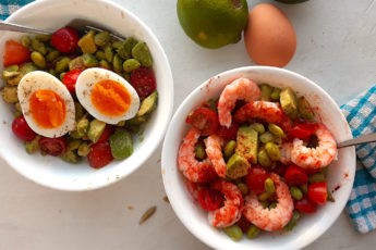 Protein Salad with edamame, sliced cherry tomatoes, avocado, eggs, and shrimp in two white bowls with silver forks on a white window sill with blue checked towels.