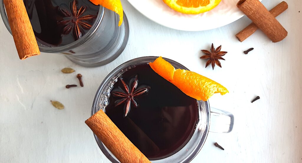 Mulled Red Wine in two clear glass mugs garnished with orange twists, star anise, and cinnamon sticks. Fresh ginger root and orange slices on a cream plate in the background. More cinnamon sticks, cardamom pods, whole cloves, and star anise scatter around on a white background. Overhead Shot. Sugar with Spice Blog.