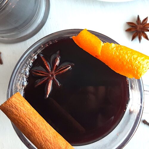 Mulled Red Wine in a clear glass mug garnished with orange twists, star anise, and cinnamon sticks. Second glass mug of mulled wine above. More cinnamon sticks, cardamom pods, whole cloves, and star anise scatter around on a white background. Overhead Shot. Sugar with Spice Blog.