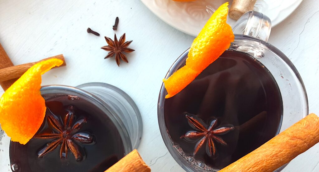 Mulled Red Wine in two clear glass mugs garnished with orange twists, star anise, and cinnamon sticks. Fresh ginger root and orange slices on a cream plate above. More cinnamon sticks, cardamom pods, whole cloves, and star anise scatter around on a white background. Overhead Shot. Sugar with Spice Blog.