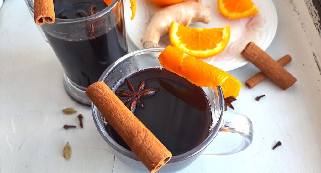 Mulled Red Wine in two clear glass mugs garnished with orange twists, star anise, and cinnamon sticks. Fresh ginger root and orange slices on a cream plate in the background. More cinnamon sticks, cardamom pods, whole cloves, and star anise scatter around on a white background. Front Shot. Sugar with Spice Blog.