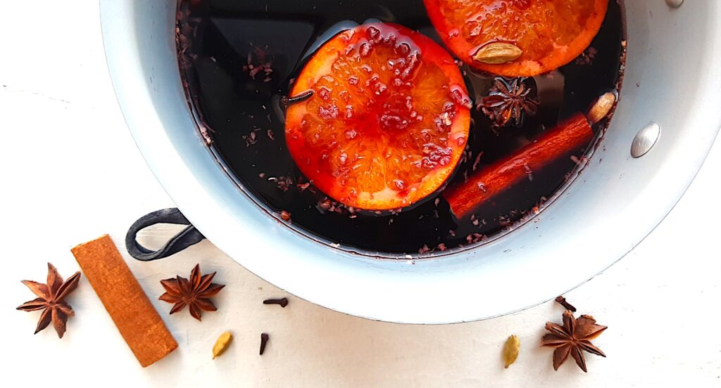 Mulled Red Wine in a sauce pan with orange rounds, cardamom pods, cinnamon sticks, cloves, and star anise. On a white background with more cinnamon sticks, star anise, cardamom pods, and whole cloves scattered around. Overhead shot. Sugar with Spice Blog.