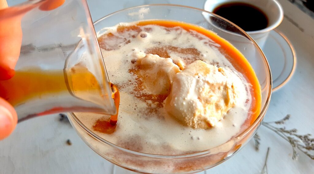 Earl Grey tea affogato preparation close up. A woman's hand pours a carafe of espresso over a glass coupe with earl grey tea ice cream. A white espresso cup with more espresso to one side. A silver spoon, lavender sprigs, and babies breath flowers scattered around on the white background. Sugar with Spice Blog.