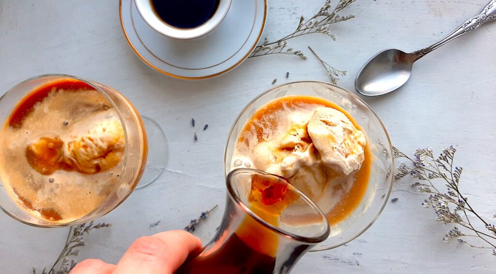 Earl Grey tea affogato preparation. A woman's hand pours a carafe of espresso over a glass coupe with earl grey tea ice cream. A second coupe with earl grey tea ice cream and espresso and a white espresso cup with more espresso to one side. A silver spoon, lavender sprigs, and babies breath flowers scattered around on the white background. Sugar with Spice Blog.