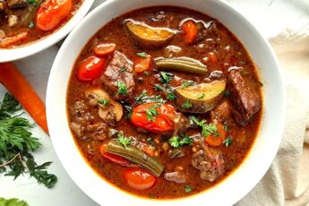 Classic stovetop beef stew with beef cubes, zucchini, carrots, tomatoes, green beans, mushrooms, and eggplant in a rich red wine broth. All served in a white bowl. A second white bowl to one side. Fresh carrots and carrot tops, a silver spoon, and a linen cloth to one side. All on a white background, overhead shot. Sugar with Spice Blog.