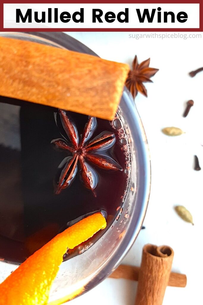 Mulled Red Wine in a clear glass mug garnished with orange twists, star anise, and cinnamon sticks. Only half the cup displayed in the photo. More cinnamon sticks, cardamom pods, whole cloves, and star anise scatter around on a white background. Overhead Shot. Text overlay with red border reads mulled red wine. Pinterest image. Sugar with Spice Blog.